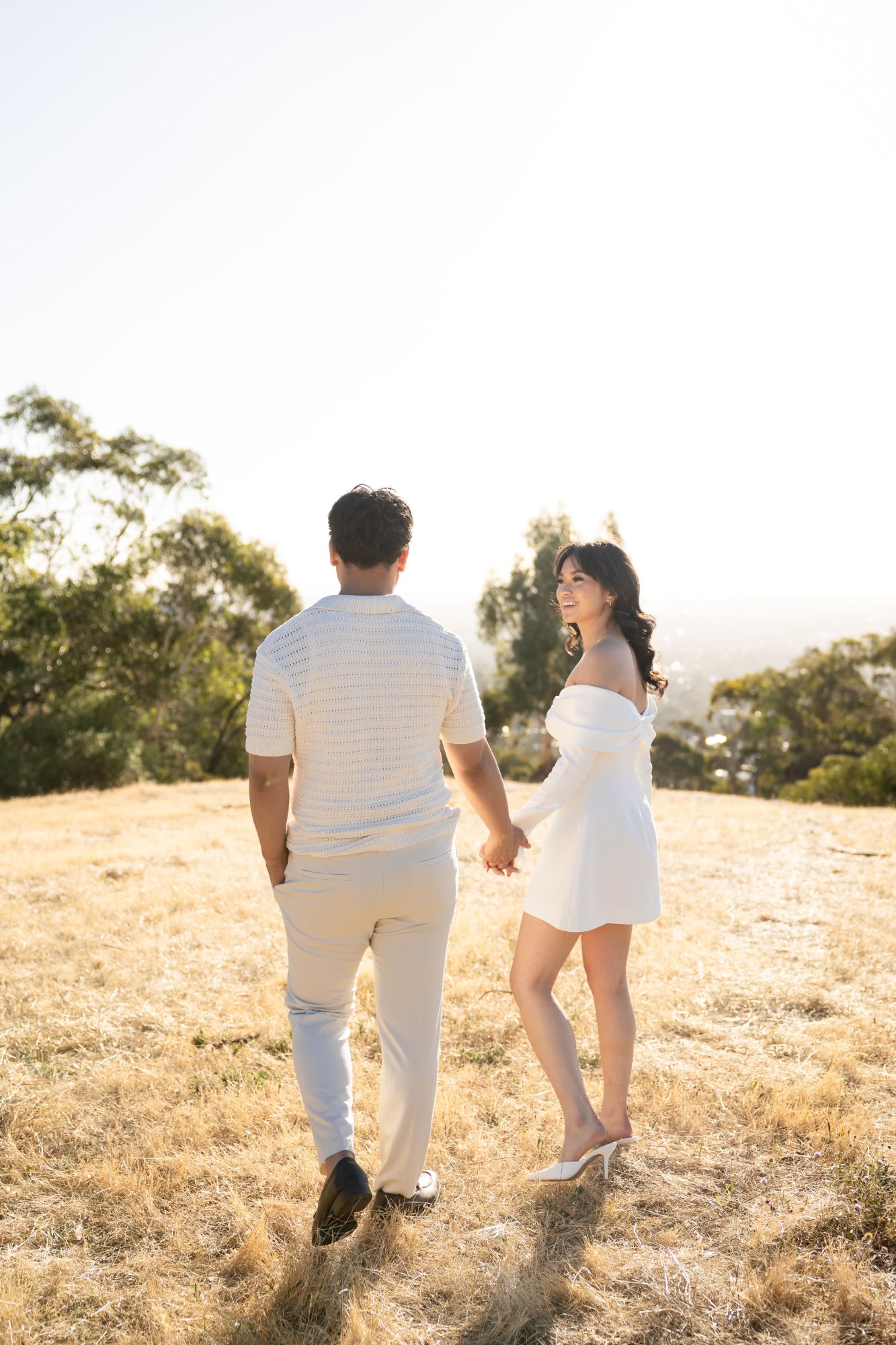 Couple walking hand in hand across golden grass as she looks back smiling