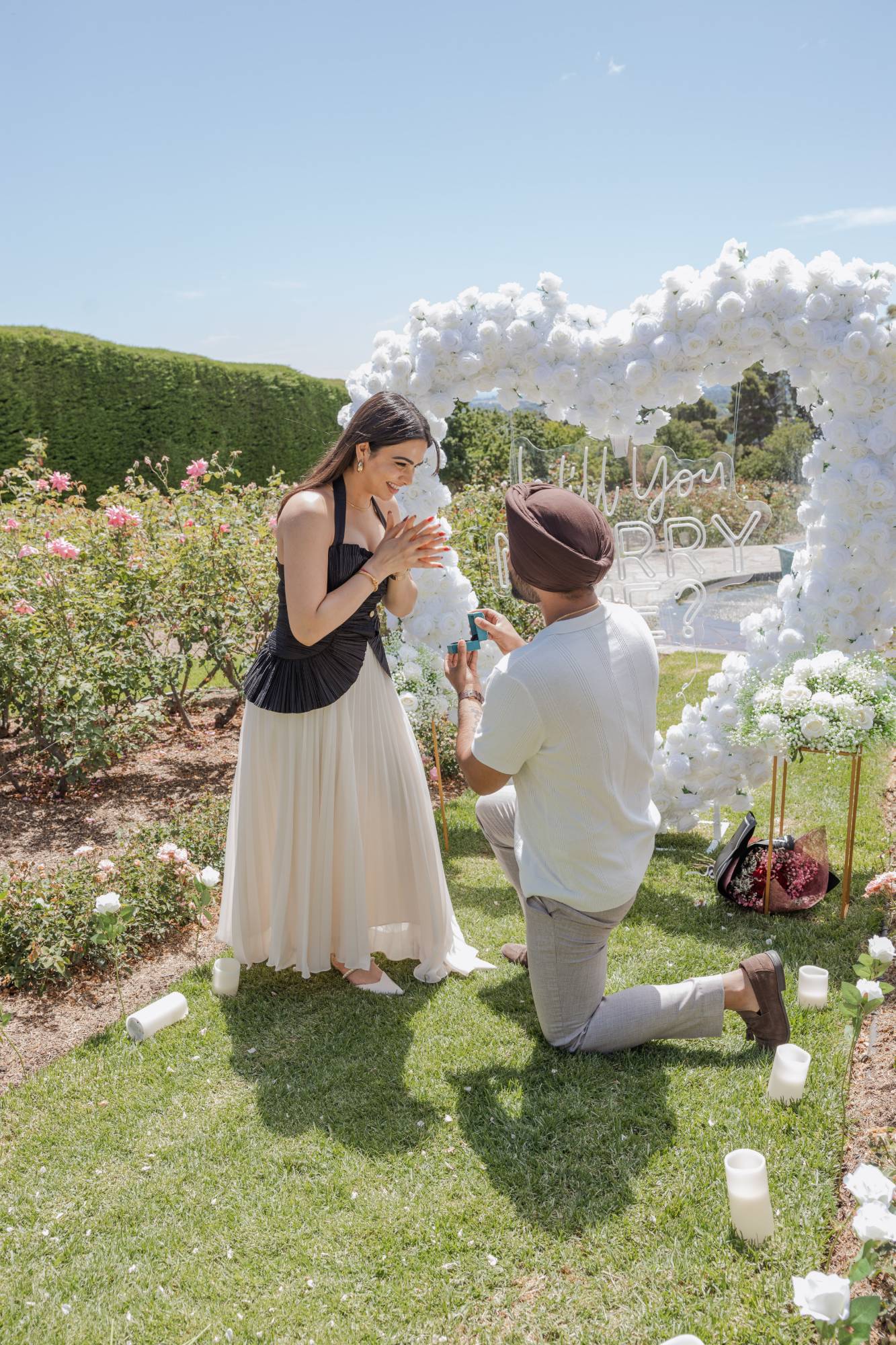 Man on one knee holding out a ring beside a white rose arch in a rose garden