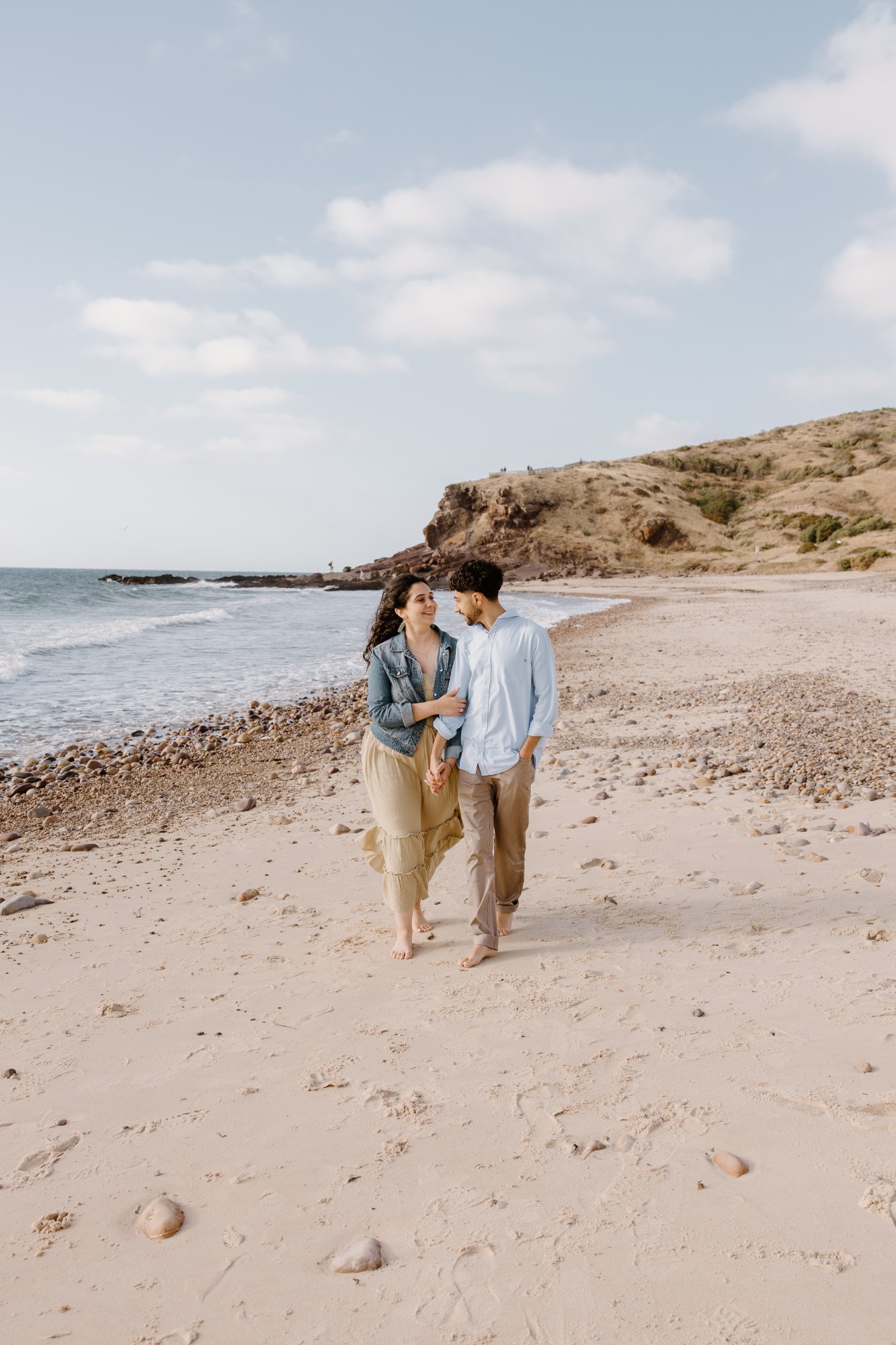 Engaged couple walking barefoot along a beach below a headland