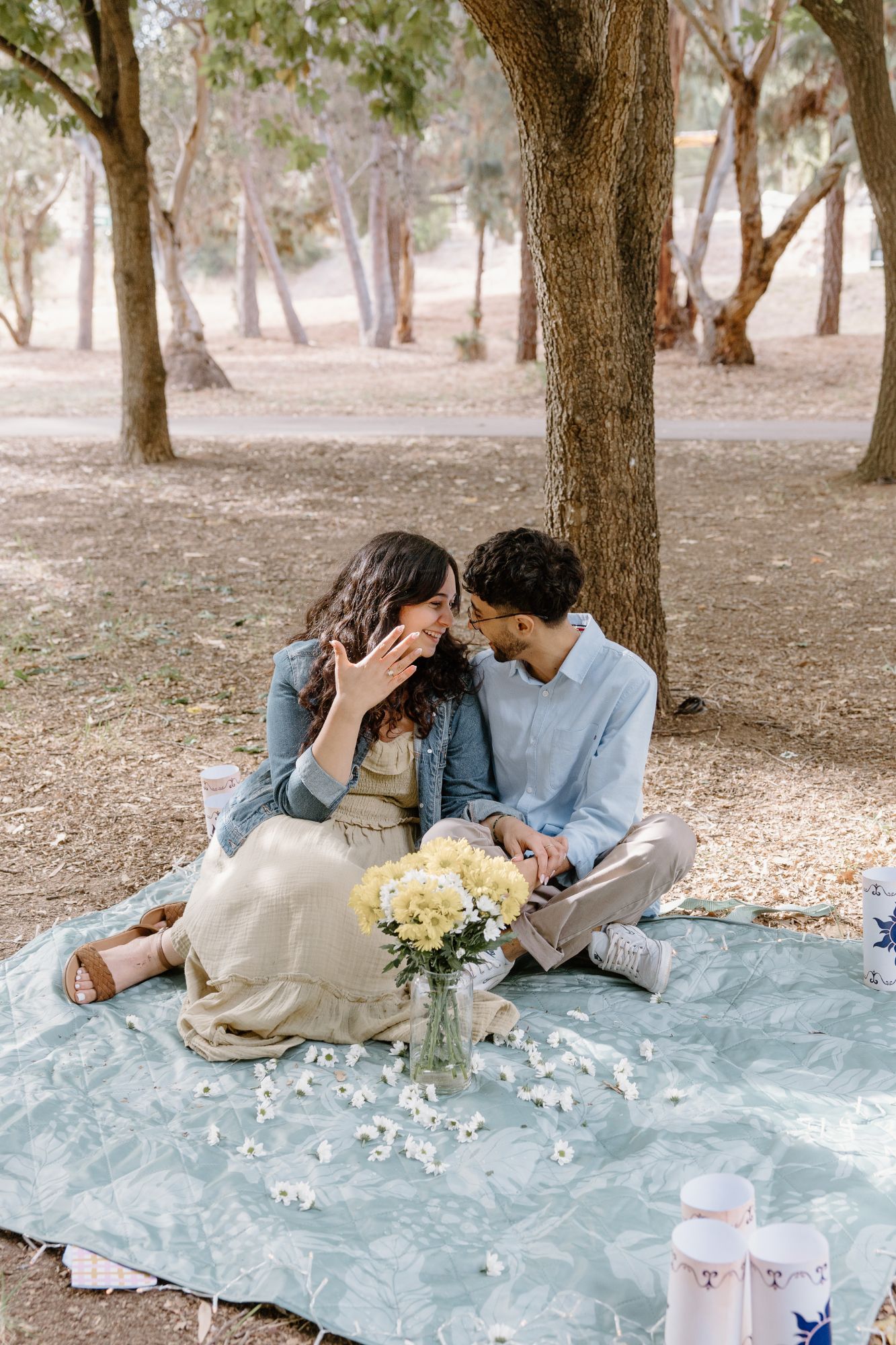 Couple on a picnic blanket under a tree as she shows off her new ring