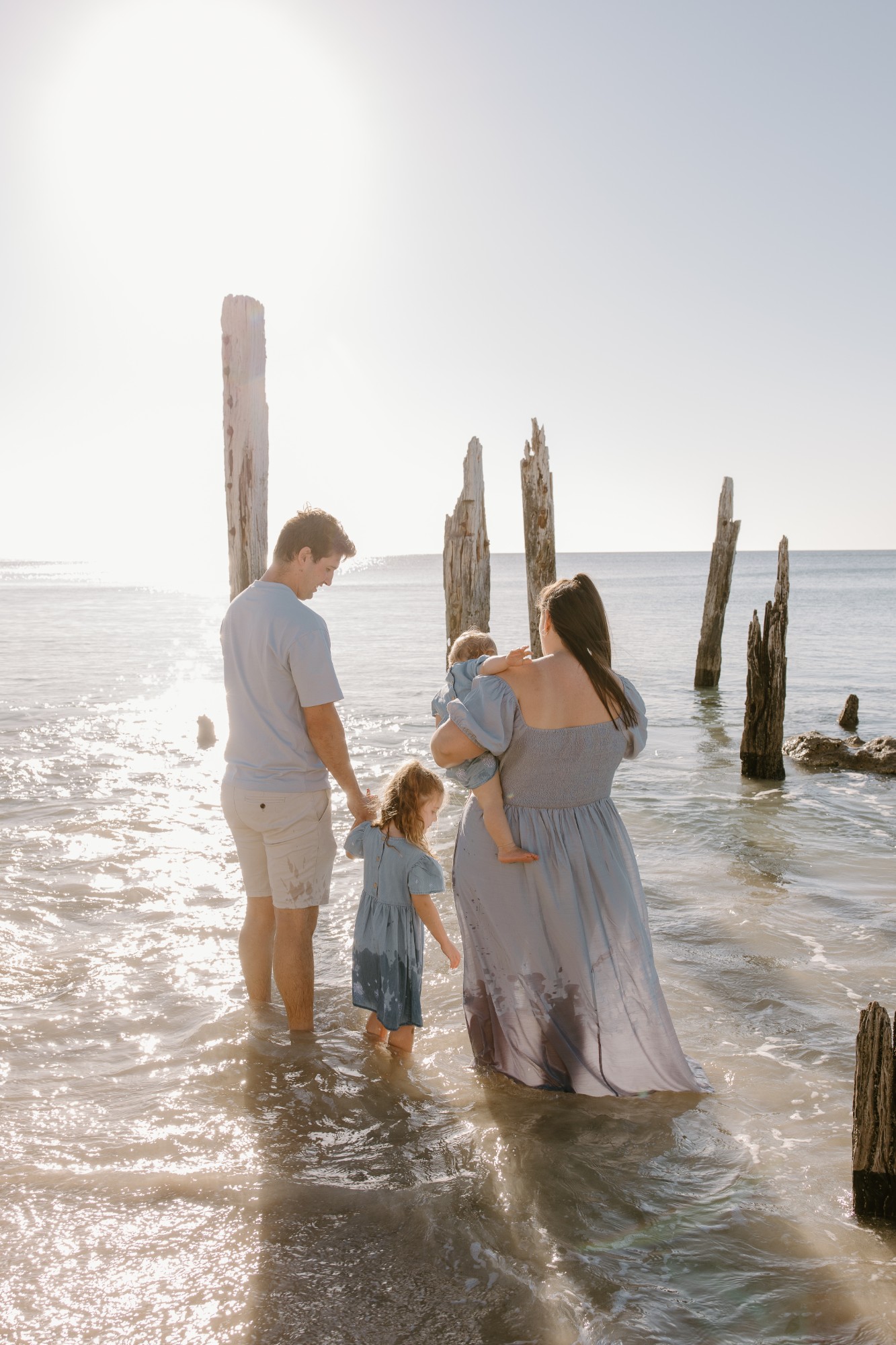 Family wading into the sea among old jetty posts, sun flaring