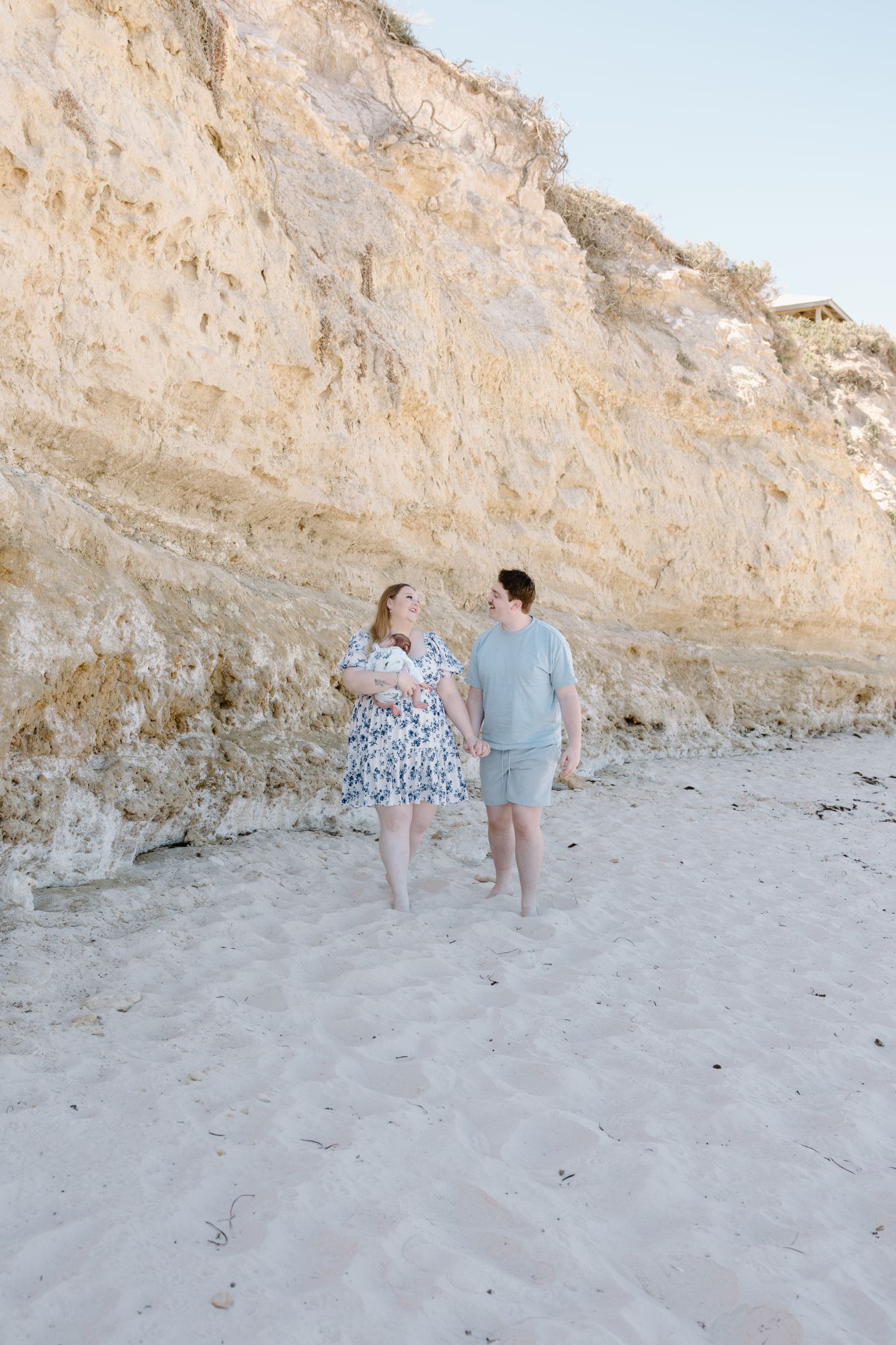 Couple walking hand in hand below a cliff, their newborn in her arms