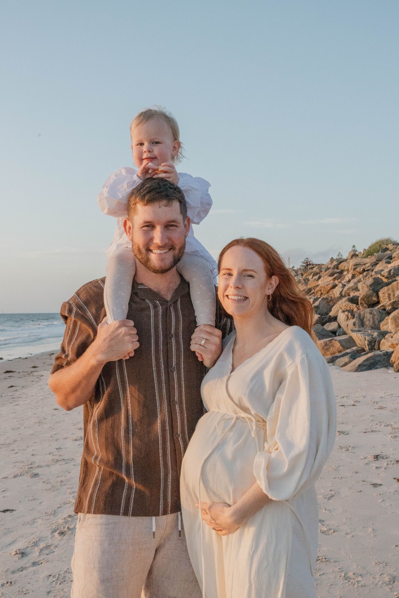 Toddler on dad’s shoulders beside his pregnant mum, the sea behind