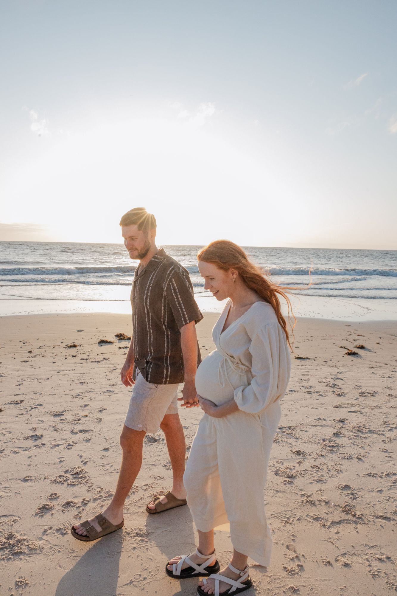 Couple walking on the beach at sunset, her hand on her bump