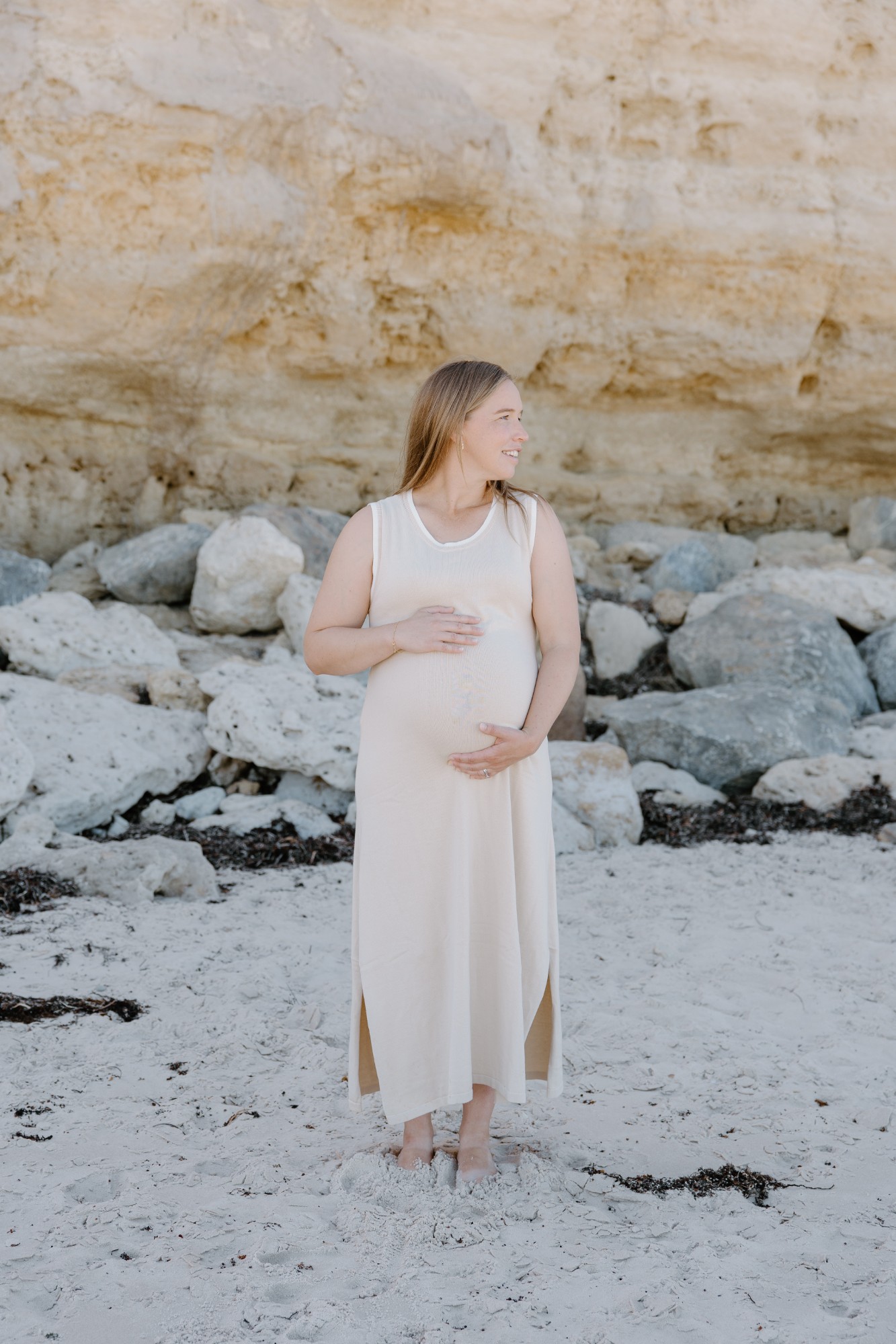 Pregnant mother in a cream dress on the sand, hands on her bump