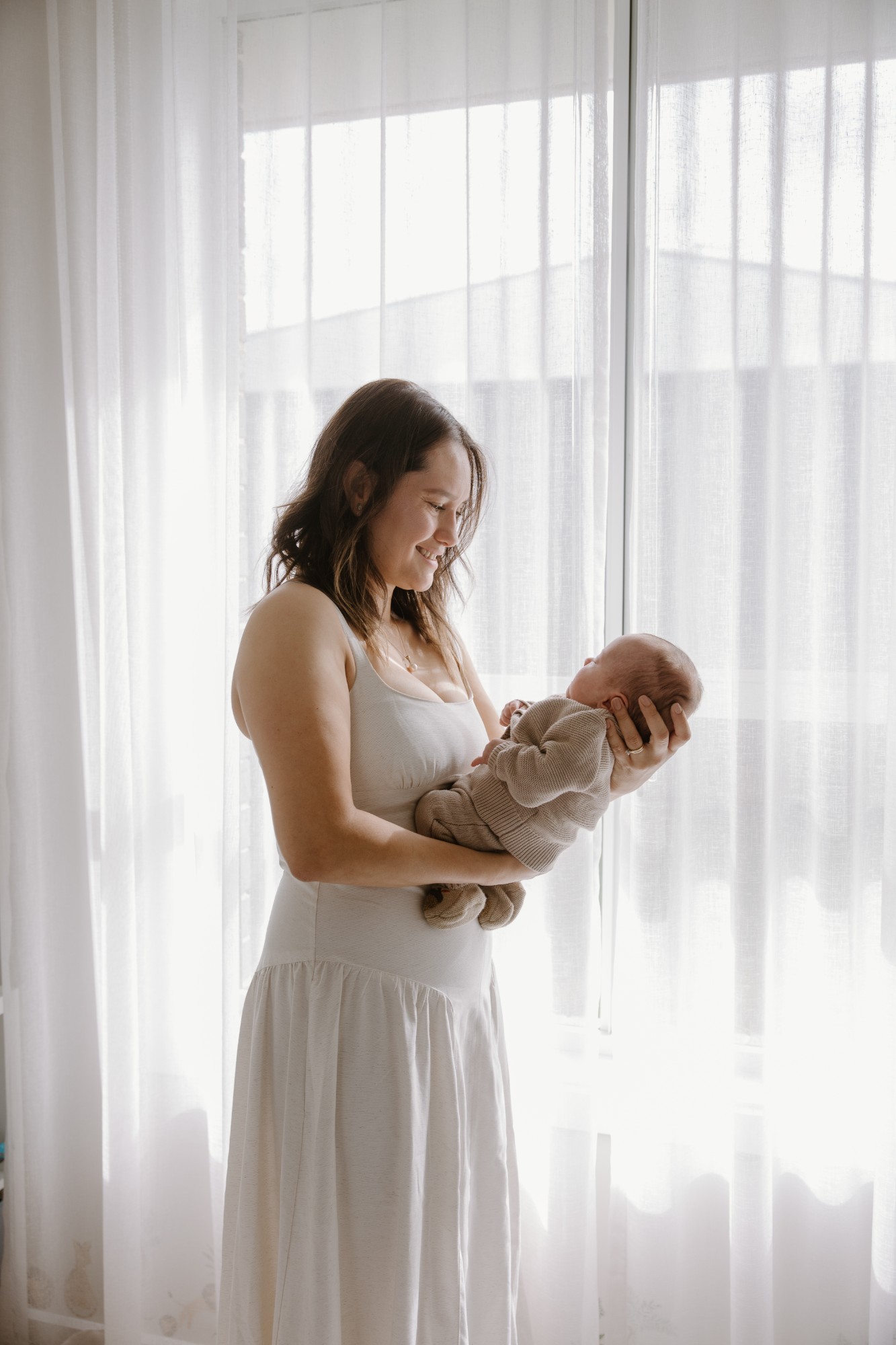 Mother holding her newborn by a sheer-curtained window