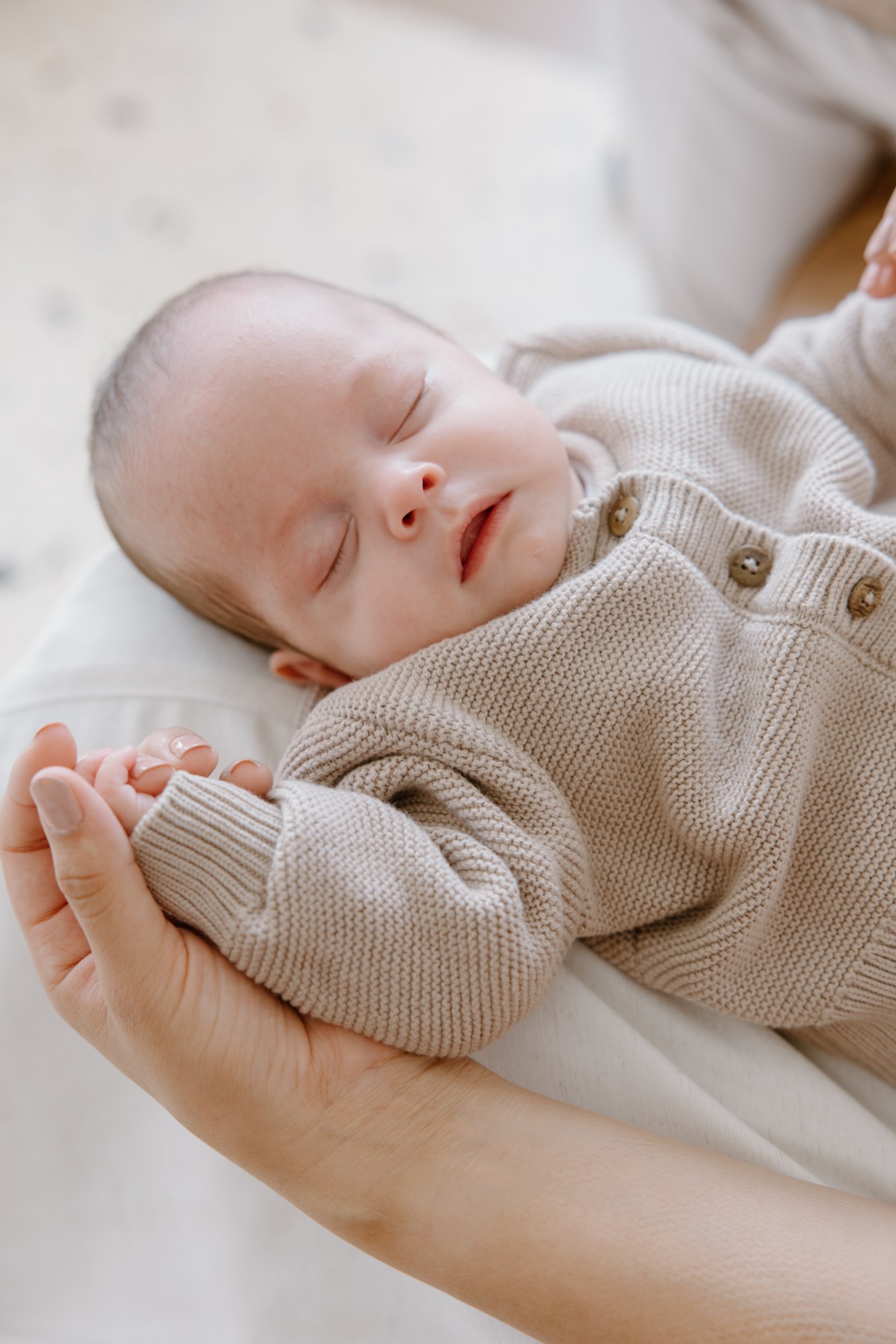 Newborn asleep in a knitted suit, his hand resting in his mother’s
