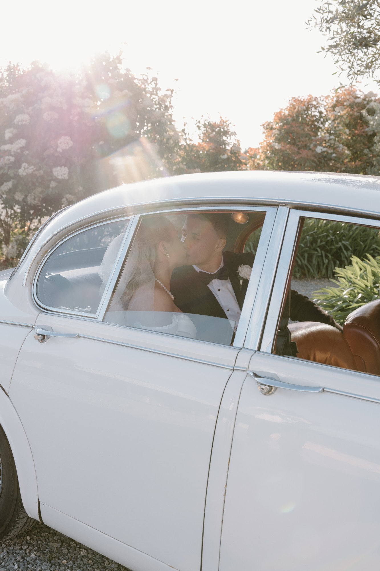 Couple kissing in the back of a white vintage Jaguar as the sun flares through