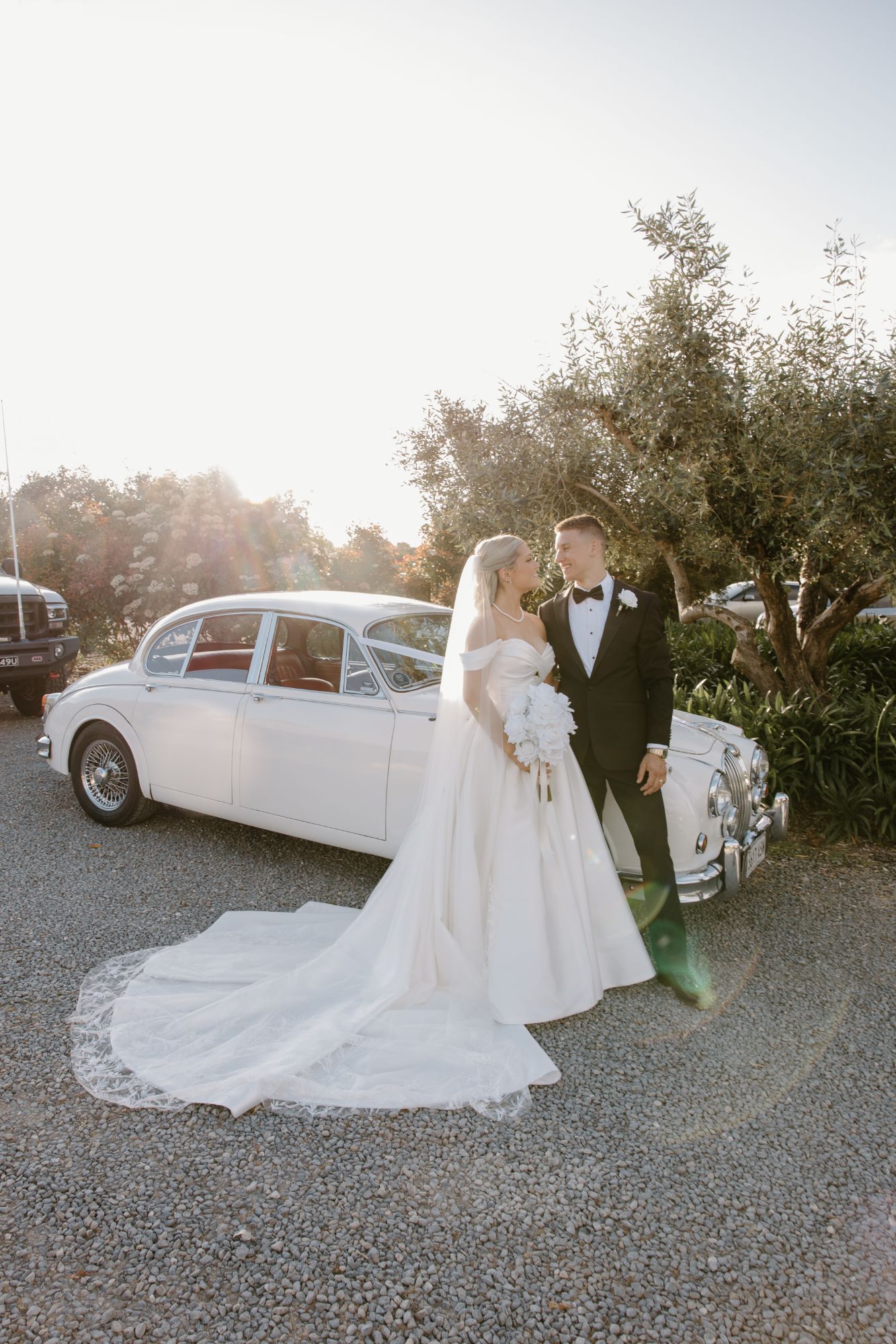 Bride and groom by the Jaguar with her long veil spread across the gravel