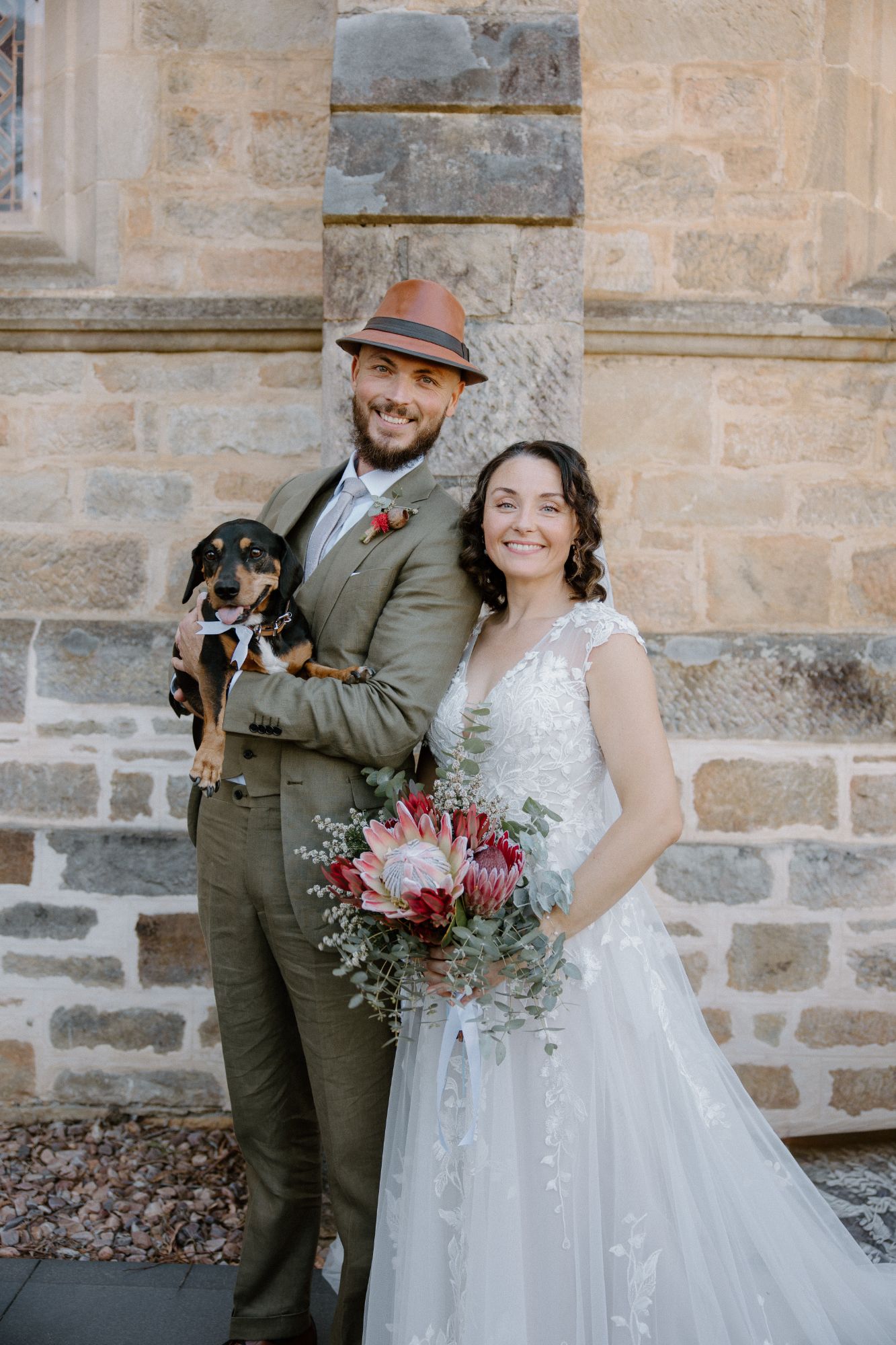 Bride and groom smiling against a sandstone wall, the groom holding their dachshund