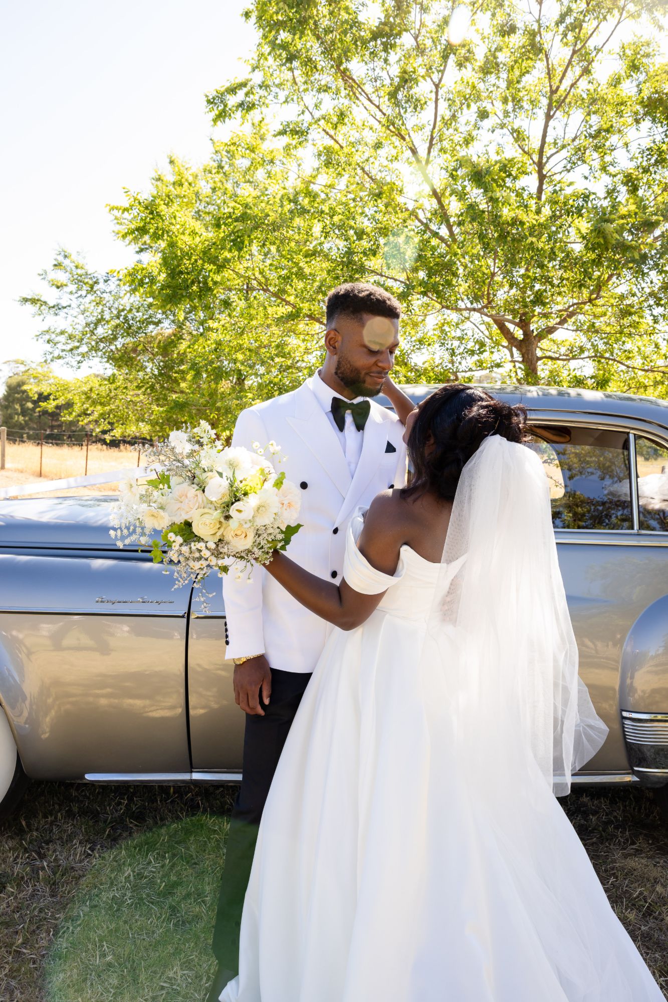 Bride holding her bouquet and touching the groom’s face beside a silver vintage car